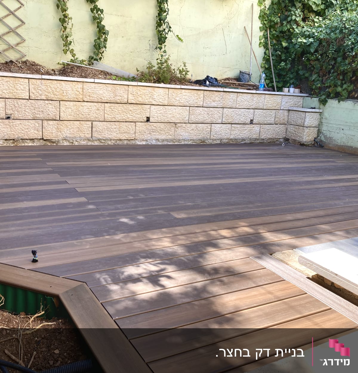 דק עץ בגינה עם קיר אבן וצמחייה מסביב